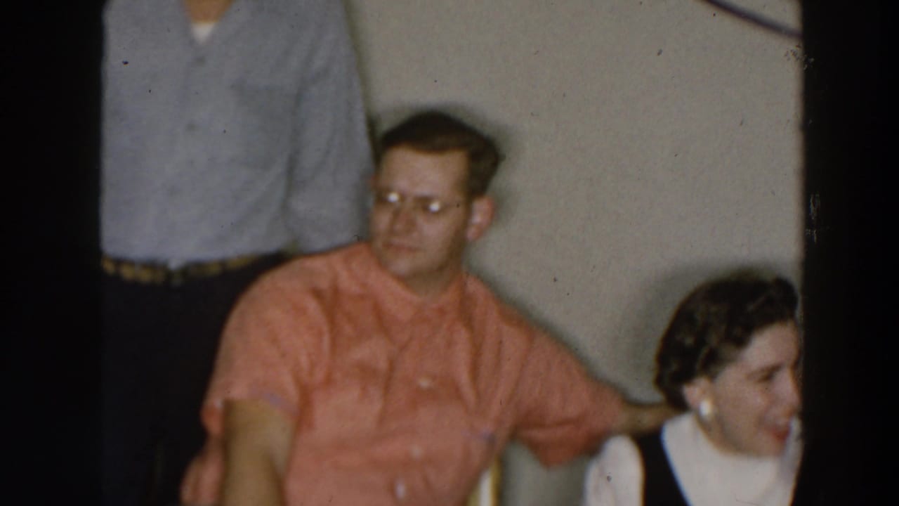Seated man in coral shirt and glasses beside a woman in a black-and-white dress, with another man standing behind them.