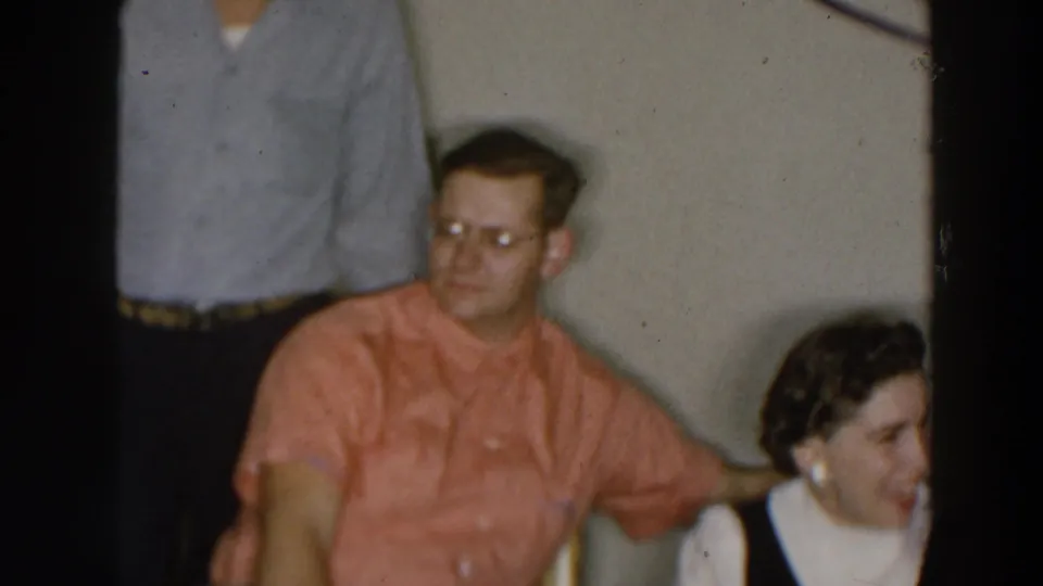 1957 Wheeling, Ohio home-movie frame showing a seated man in a coral shirt and glasses beside a woman in a black-and-white dress indoors.
