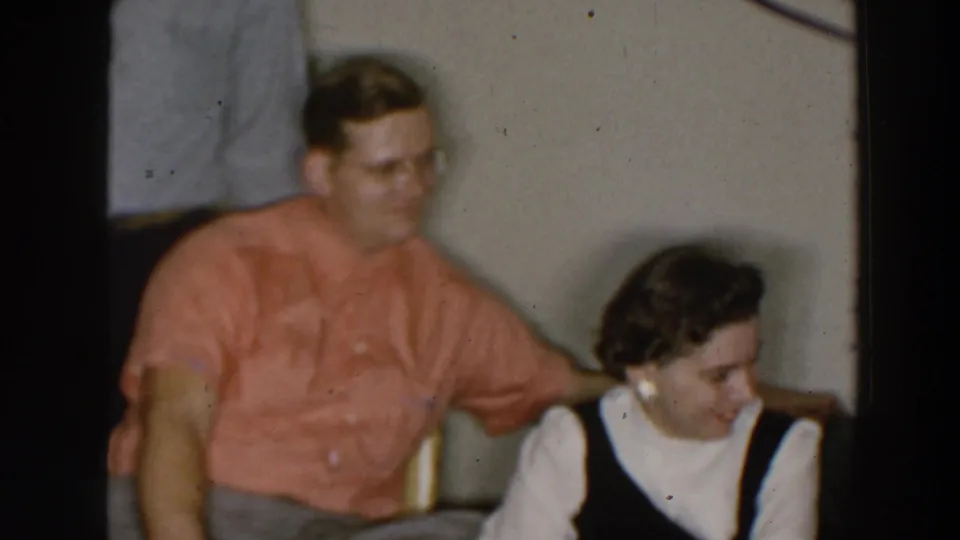1957 Wheeling, Ohio frame of adults seated indoors, with a man in a coral shirt beside a woman wearing pearl earrings and a dark dress.