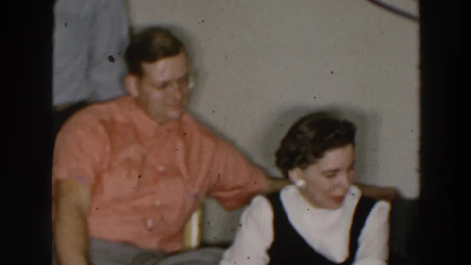1957 Wheeling, Ohio home-movie frame showing a seated man and woman sharing a quiet indoor social moment.