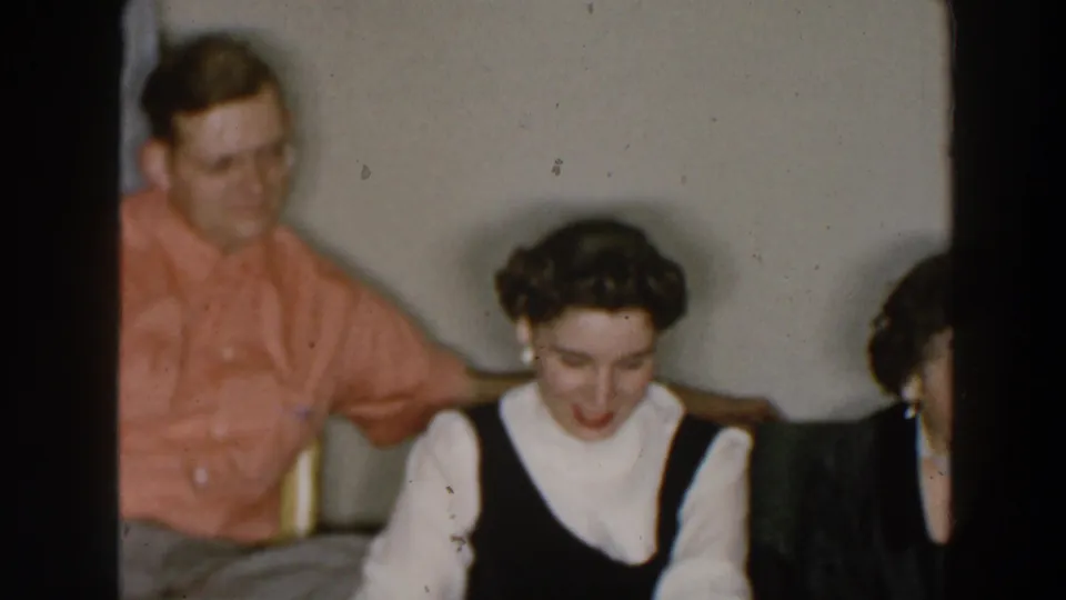 1957 Wheeling, Ohio frame with adults seated in a private interior, including a woman looking down and a man in a coral shirt nearby.