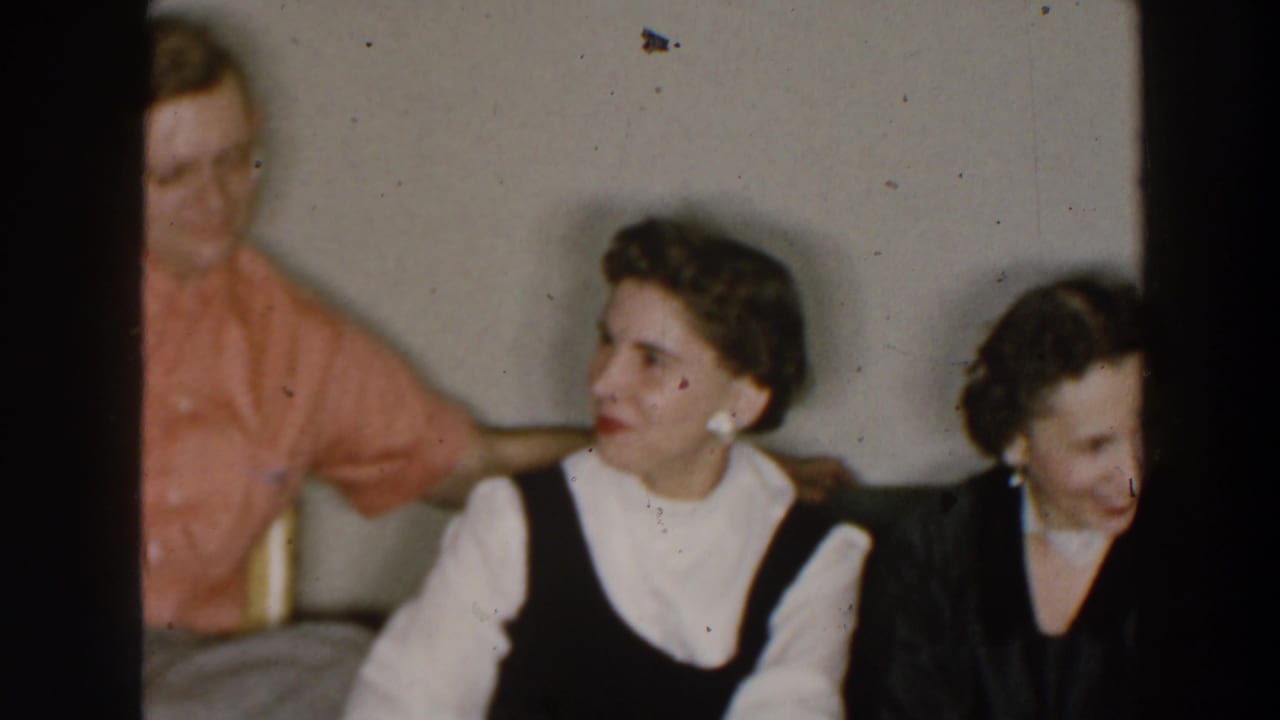 1957 8mm home-movie frame identified as Wheeling, Ohio, showing two seated women smiling while a man in a pink shirt is partly visible behind them.
