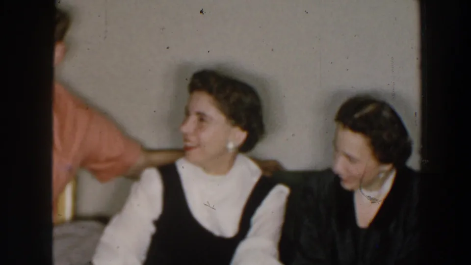 1957 home-movie frame from metadata-identified Wheeling, Ohio, with two women in dressy clothing seated close together in a private interior.