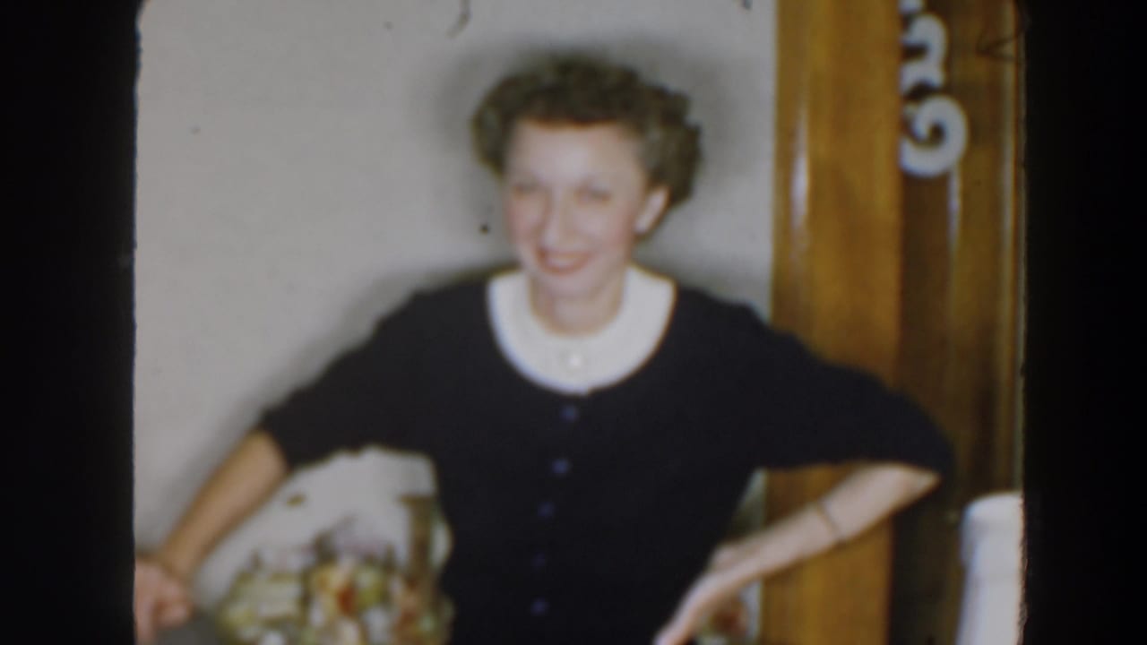 In 1957 Wheeling, Ohio home-movie footage, an adult woman smiles in a dark cardigan with a white collar near interior wood trim.
