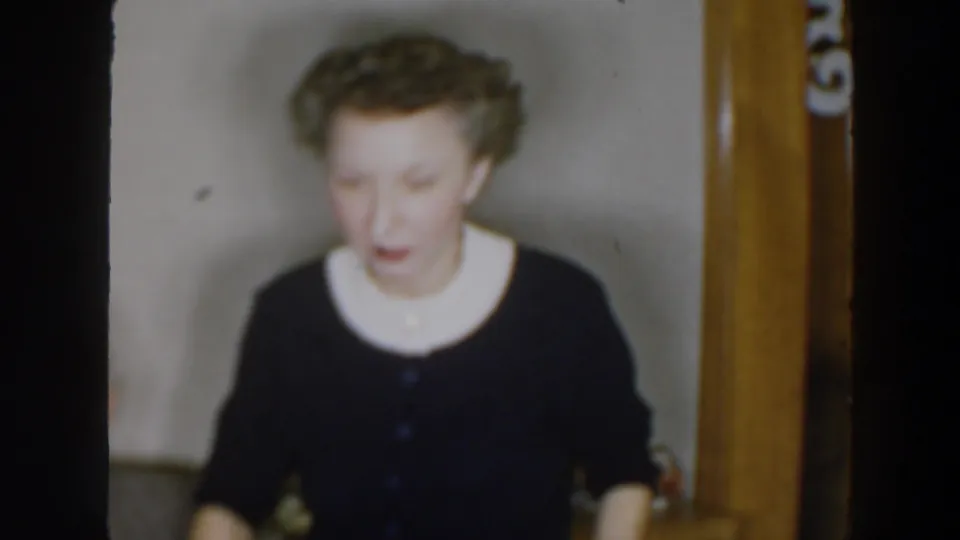 The woman faces the camera area in a softly focused 1957 8mm interior shot, with wall and wood trim behind her.
