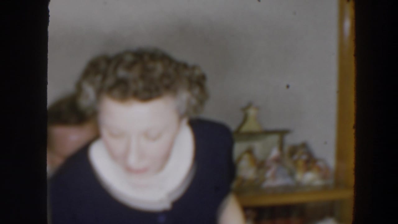 Woman in a dark dress bends forward in a living room with a partly visible man behind her.