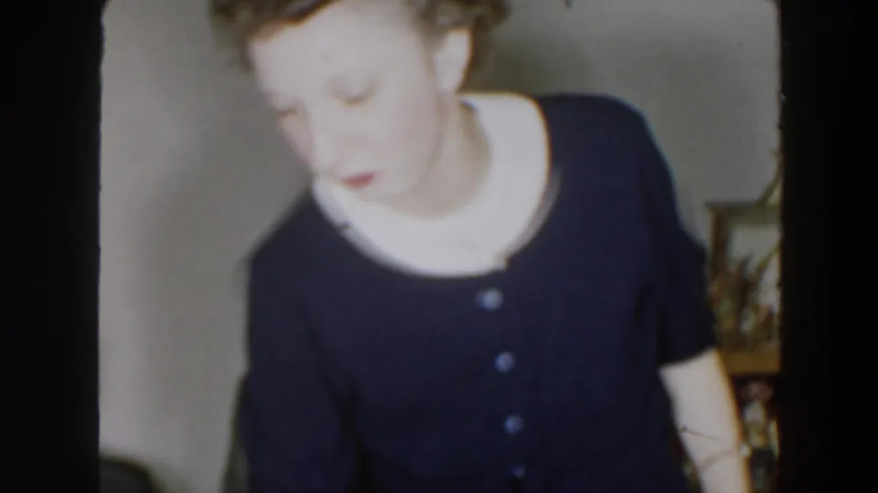 A soft 8mm frame shows the woman turning in the living room, with mid-century decor and shelving visible behind her.