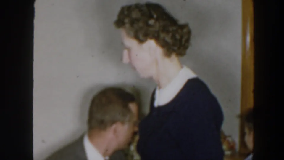 In the final frame, the woman stands in profile near a seated man, with shelving, framed decor, and a private living room setting visible.
