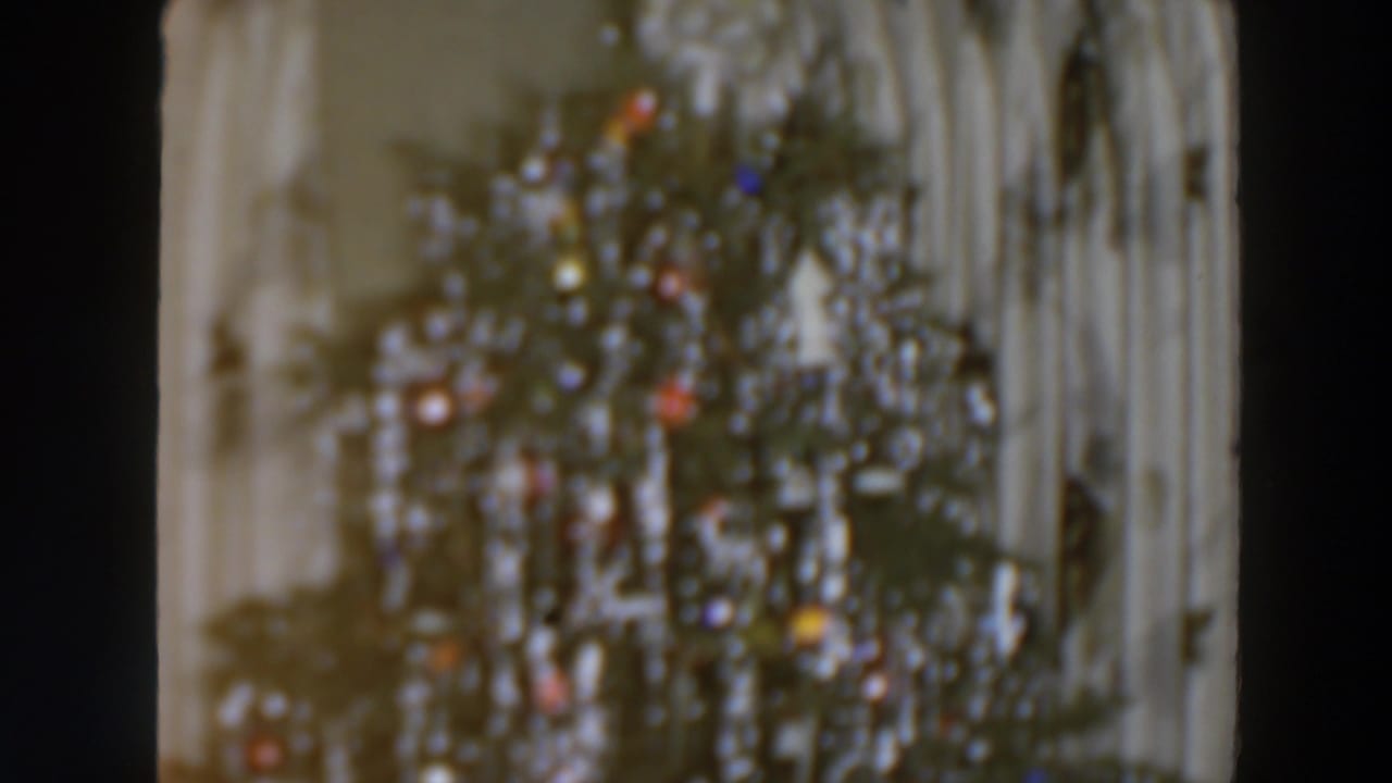 1957 home-movie frame of a tinsel-covered Christmas tree with colored ornaments or lights in front of pale curtains, likely indoors.