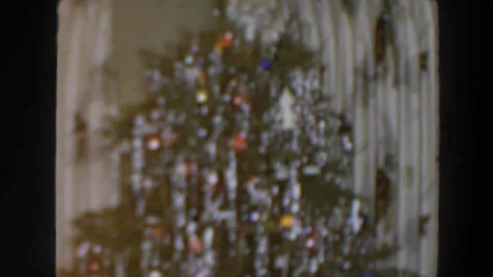 1957 home-movie frame of a tinsel-covered Christmas tree with colored ornaments or lights in front of pale curtains, likely indoors.