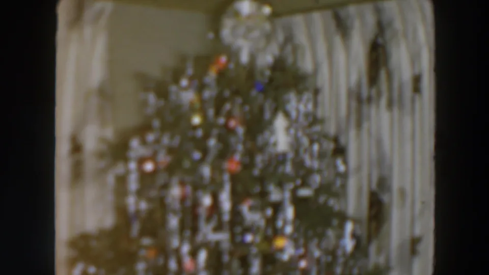 1957 8mm frame showing a Christmas tree covered in silver tinsel, colored bulbs or ornaments, and pale curtains behind it.