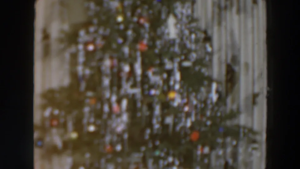 1957 8mm frame of a domestic Christmas tree with silver tinsel, colored decorations, and a pale curtained background.
