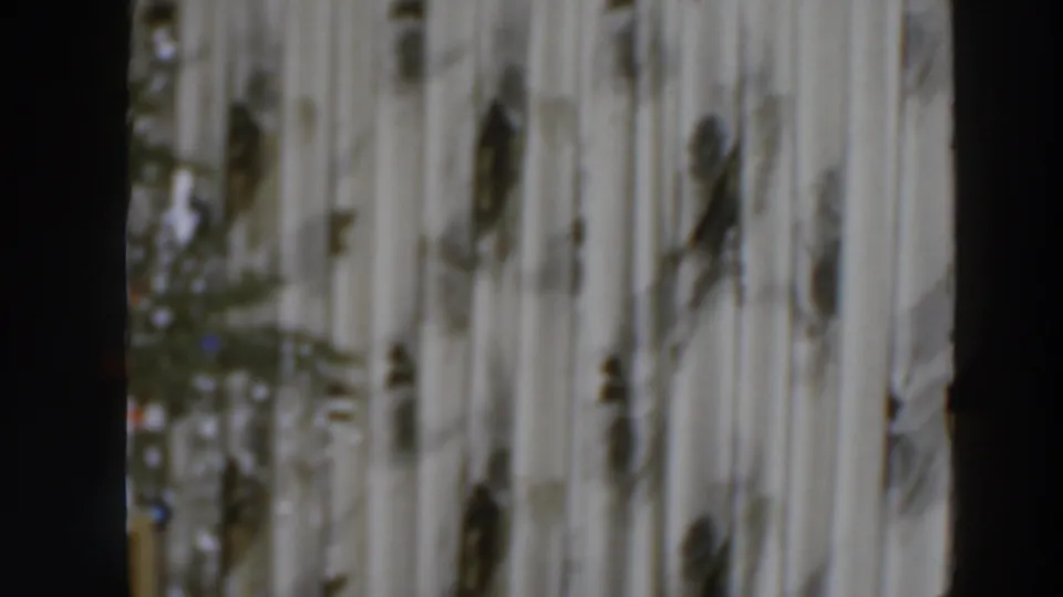 Soft 1957 8mm home-movie frame from Wheeling showing patterned curtains and a dark interior edge before the woman enters view.