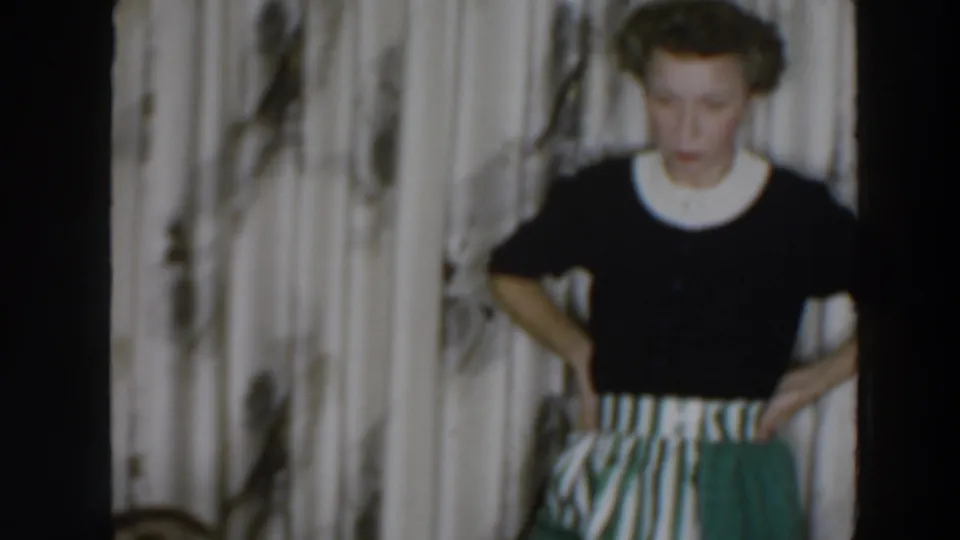 In the 1957 Wheeling home movie, the woman faces forward with hands at her waist against patterned curtains.