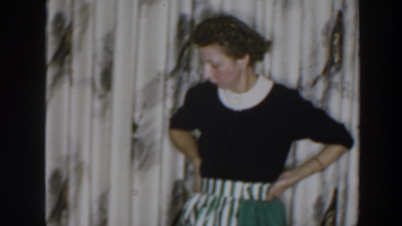 Woman in a dark top and striped skirt stands with hands on hips before patterned curtains.
