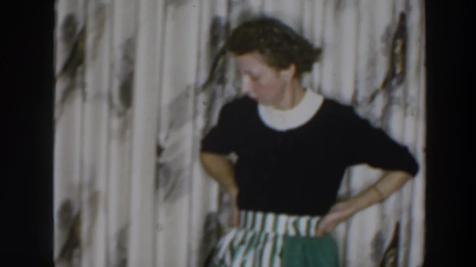 In a 1957 Wheeling, Ohio home-movie frame, a woman poses indoors with hands on hips before patterned curtains.