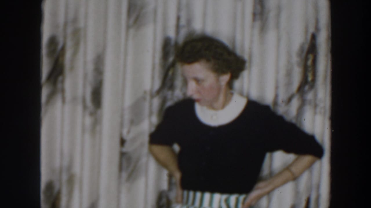 Woman stands with hands on hips in front of patterned curtains, looking down and to the side.