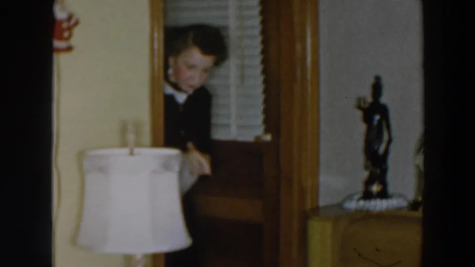 Inside the Wheeling home interior, the woman bends near a doorway beside a lamp, figurine, blinds, and wood-trimmed wall.