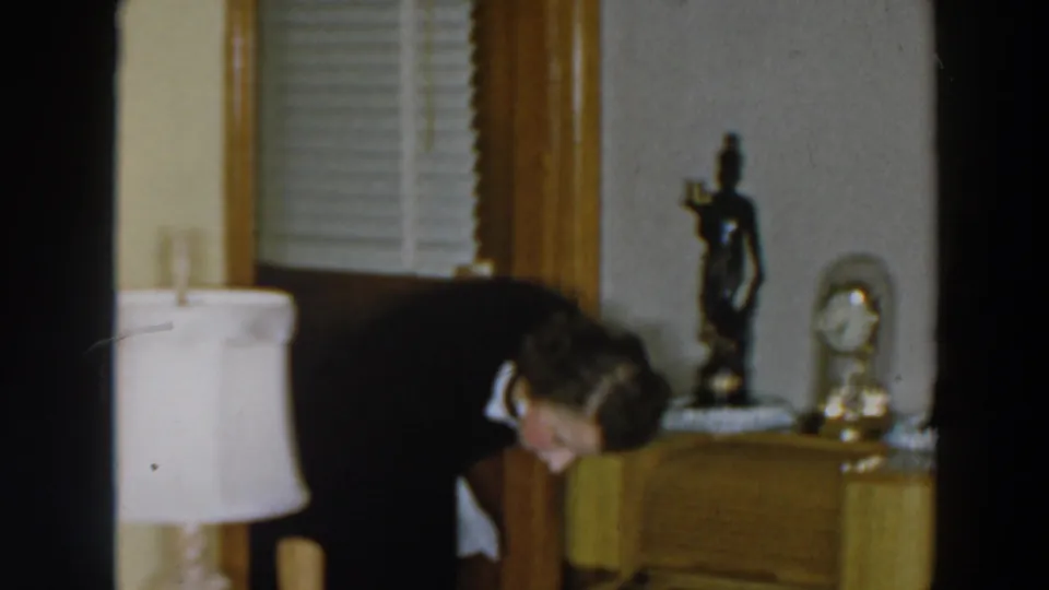 The woman bends toward the floor in the 1957 home interior, with a lamp, dark figurine, small clock, blinds, and side table visible.