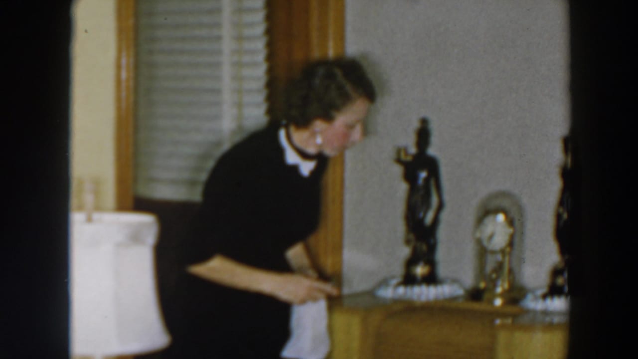 Woman in a dark dress leans toward drawers beside a lamp and tabletop decorations.