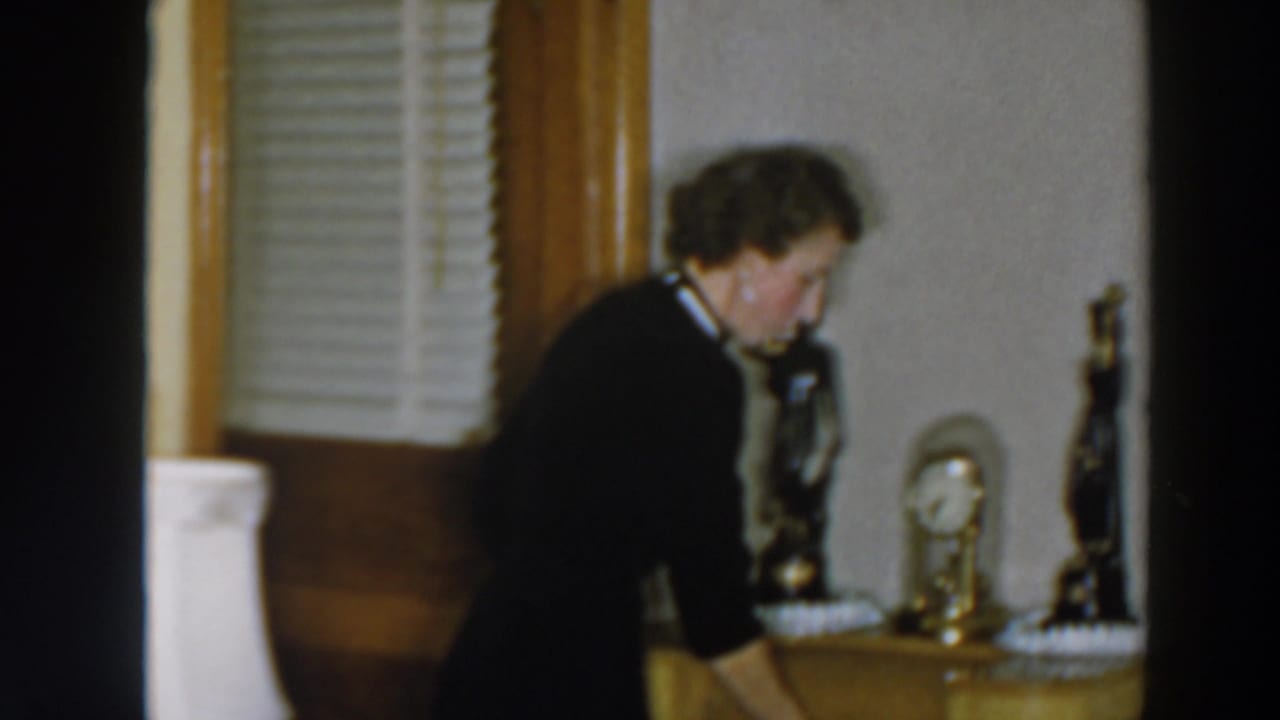 The woman bends forward in the Wheeling, Ohio interior, searching near a side table or drawers with a small clock and decorative statuettes visible.