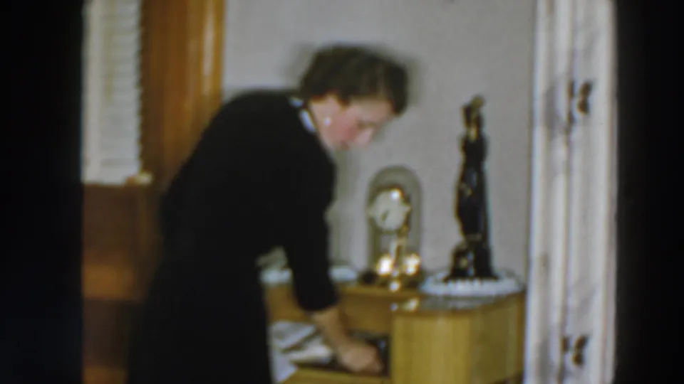 The woman reaches down toward a drawer or cabinet area in the 1957 home interior, with a small clock, statuette, wood trim, and curtains visible.