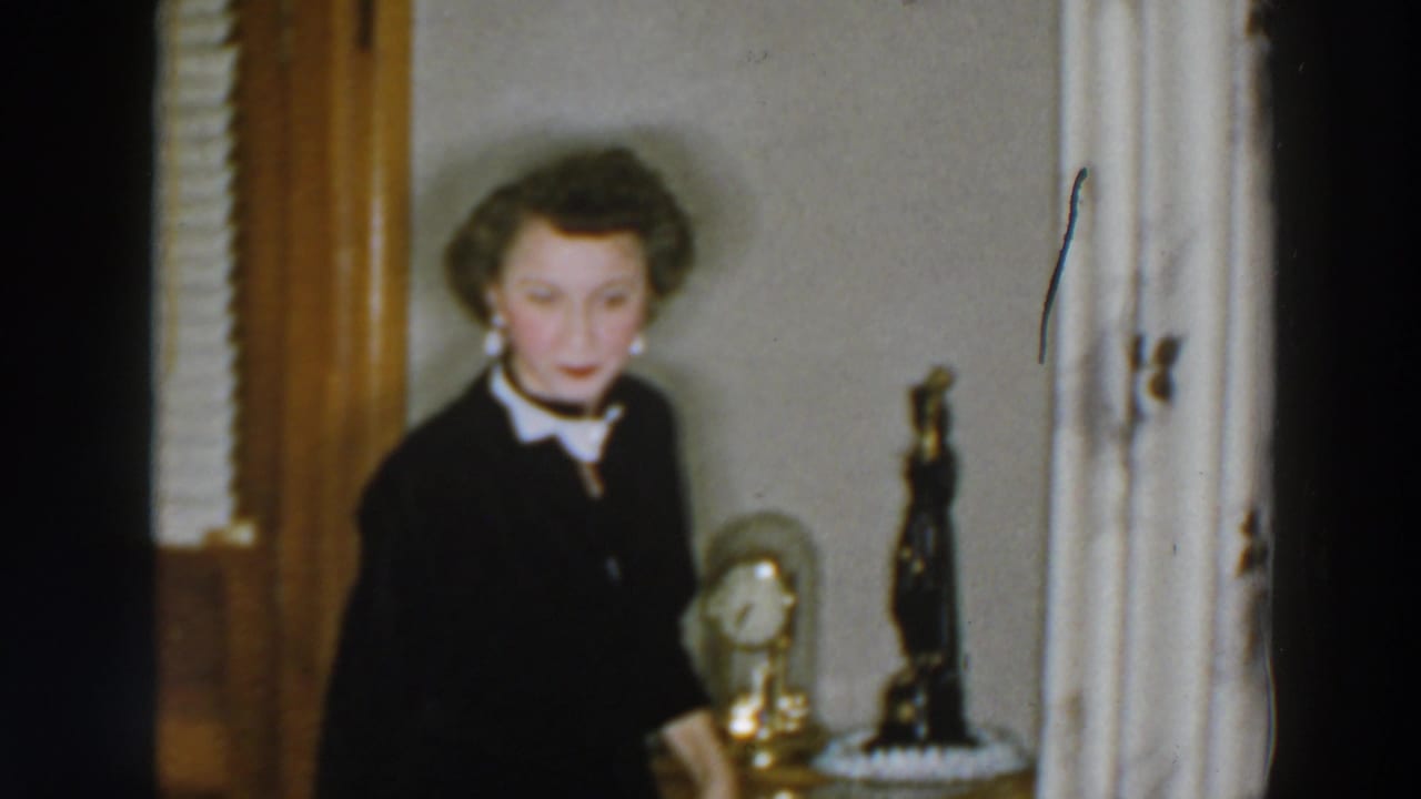 In a 1957 Wheeling home interior, a woman in a dark dress with white collar stands near a clock, statuette, wood trim, and curtains.