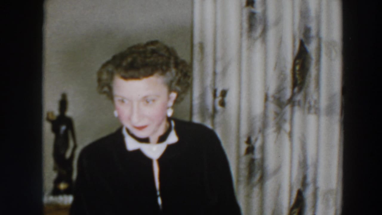 Woman faces the camera near patterned curtains and a tabletop statuette.