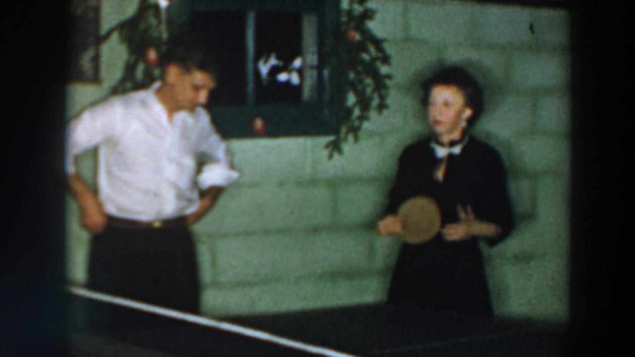 Two adults stand by a ping pong table in an indoor room with holiday greenery.
