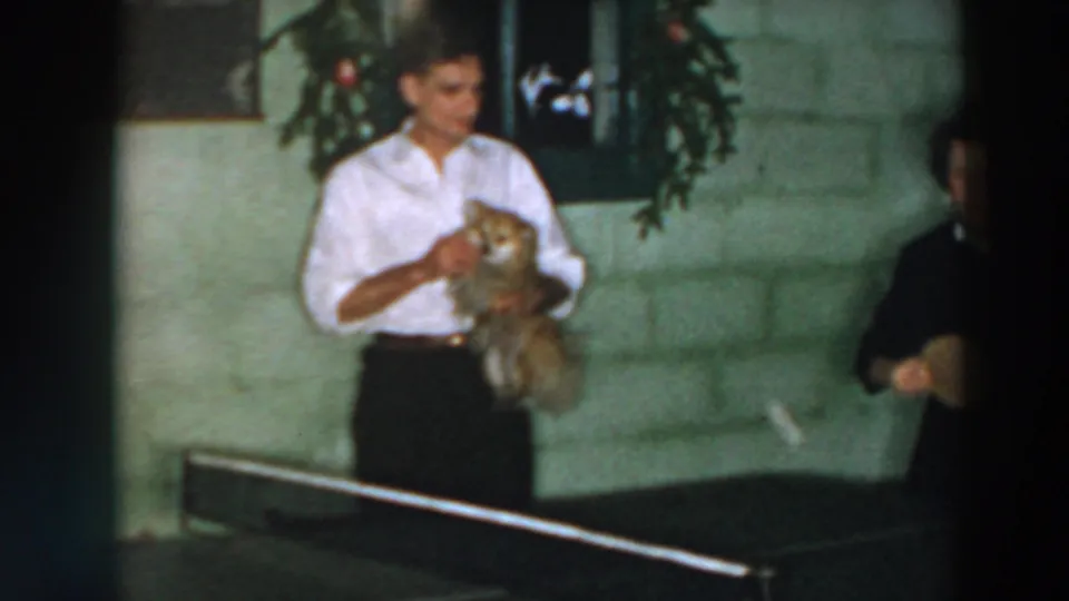 In 1957 Wheeling, Ohio home-movie footage, a man holds a small dog near a ping pong table in a concrete-block room.