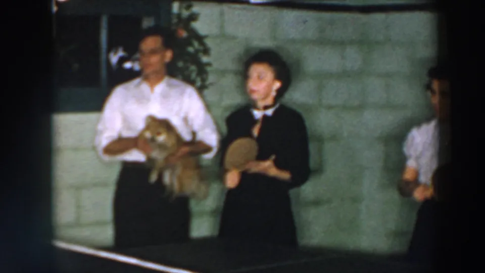 A man holding a small dog stands next to a woman with a paddle in the 1957 Wheeling, Ohio ping pong scene.