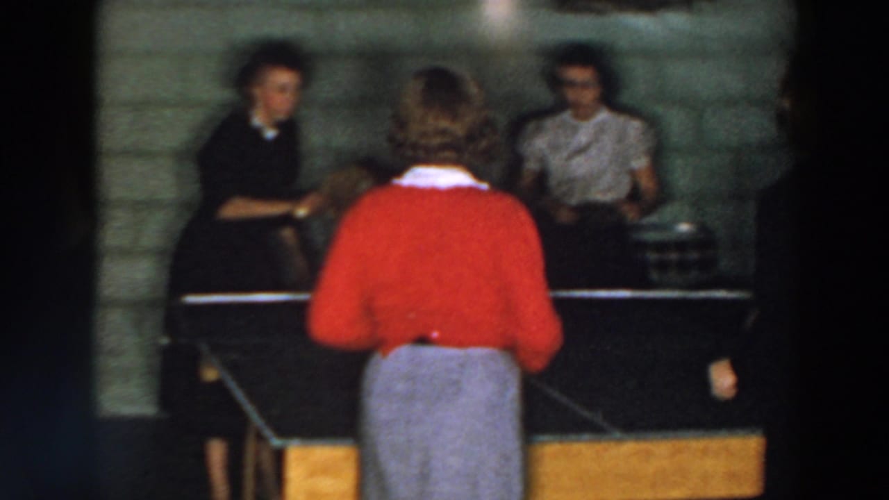 1957 Wheeling, Ohio home-movie frame showing women around a ping pong table in a cinder block interior.