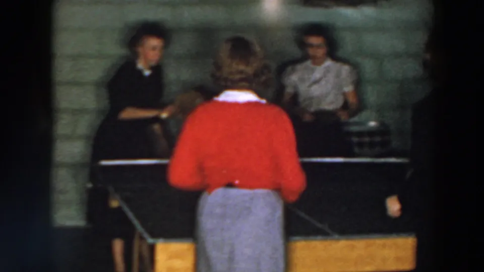 1957 Wheeling, Ohio home-movie frame showing women around a ping pong table in a cinder block interior.