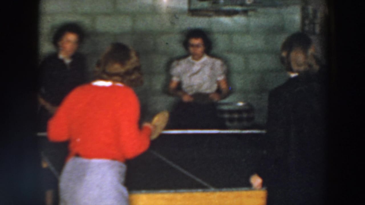 A woman in a red sweater plays table tennis while others watch nearby.
