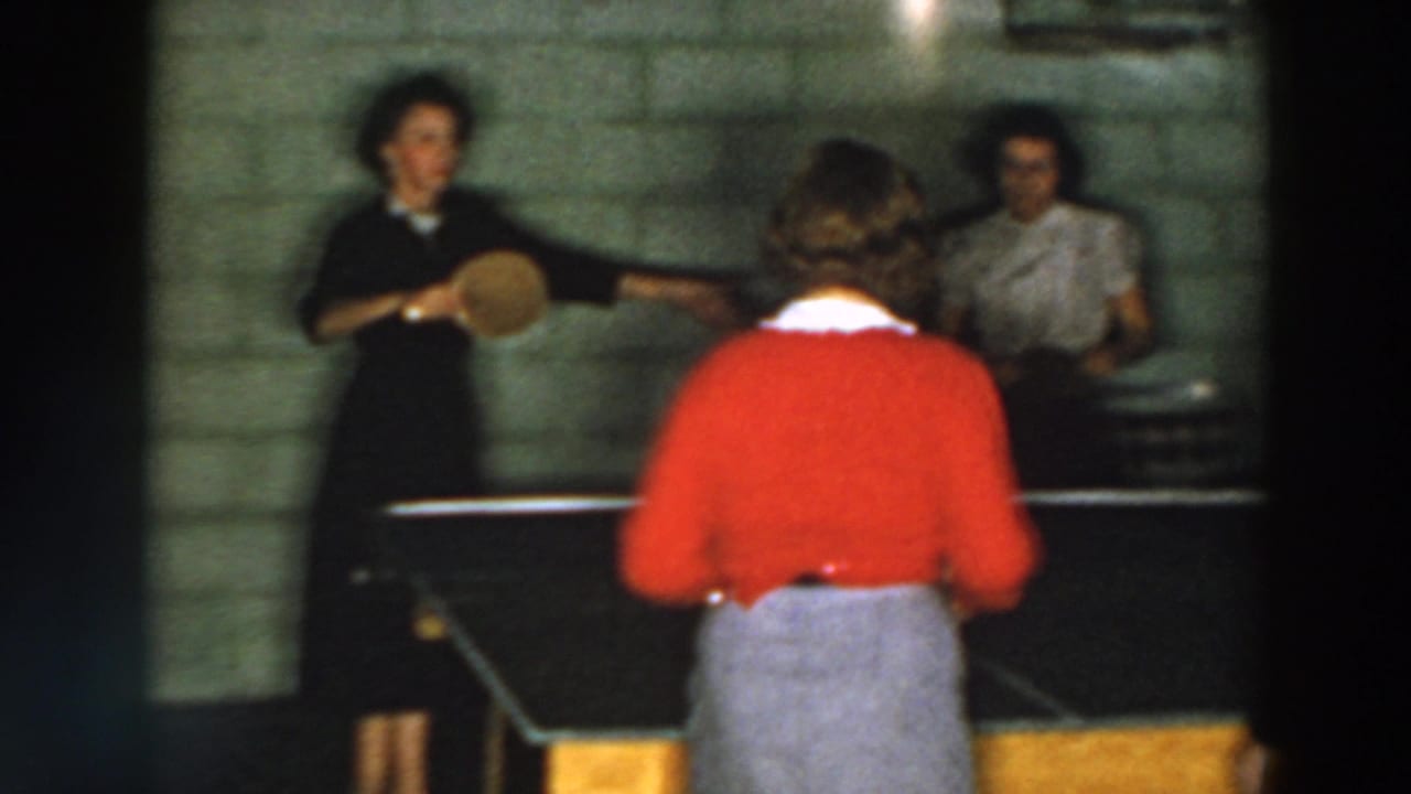 Three women play table tennis in a dim basement with a cinder block wall.