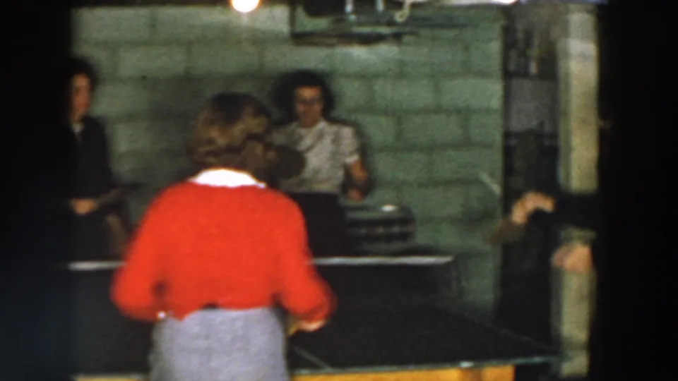 1950s home-movie frame of women continuing a table tennis game in a dim cinder block basement in Wheeling, Ohio.