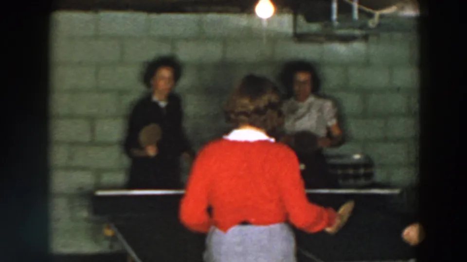 Wheeling, Ohio home-movie frame showing women at a basement table tennis table under warm overhead light, with soft 8mm blur.