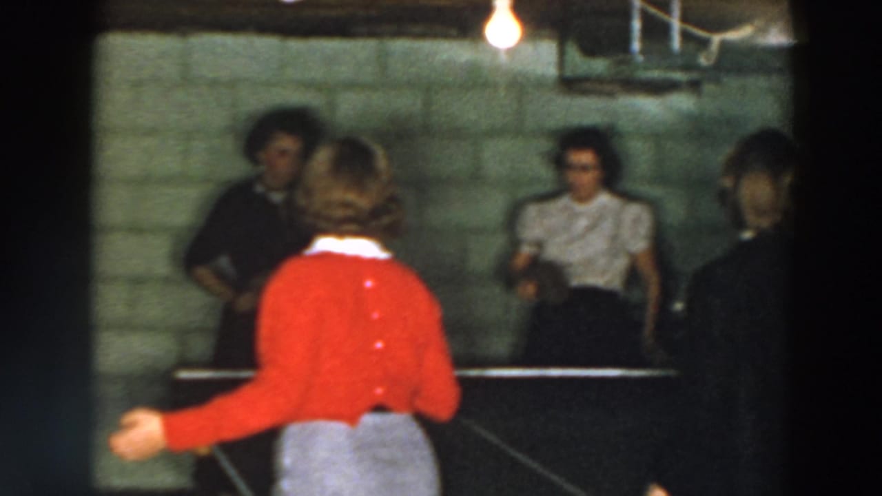 Woman in a red cardigan reaches during a basement table tennis game with other women watching.