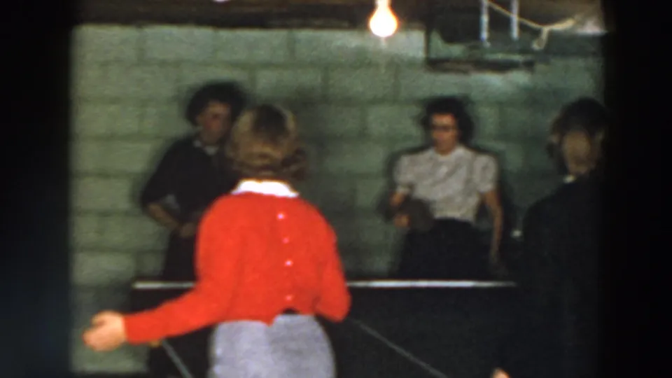 A 1957 Wheeling, Ohio basement scene shows a woman in red extending her arm at a ping pong table while other women stand beyond it.