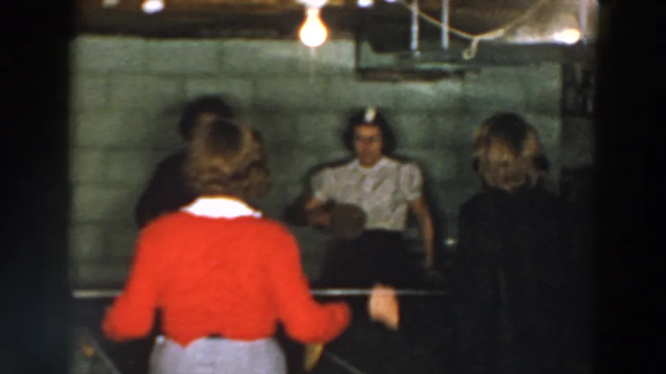 The home-movie frame shows the red-cardigan player from behind, a seated woman across the table, and another woman at the side in a basement setting.