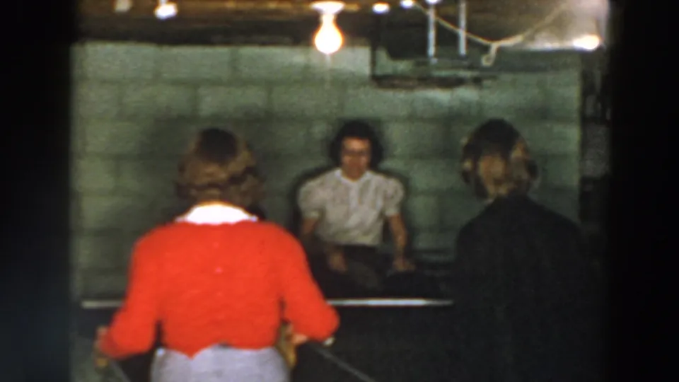A soft 8mm frame from Wheeling, Ohio shows women around a basement ping pong table, with a red cardigan foreground and cinder block wall behind.