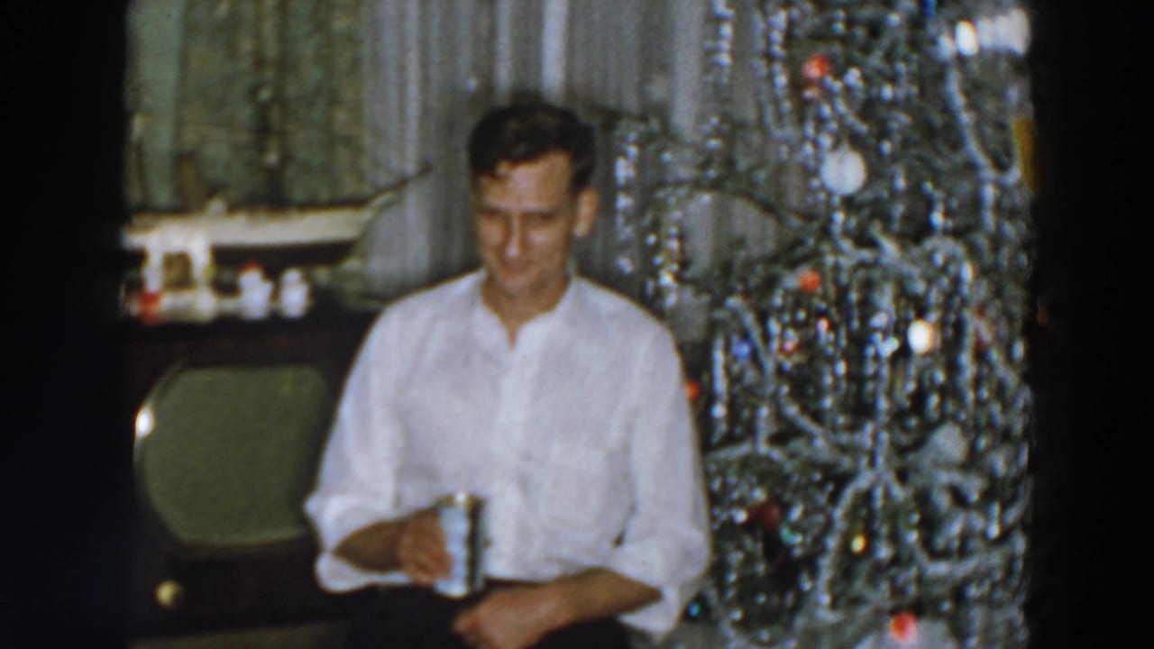 Man in a white shirt seated beside a tinsel Christmas tree in a living room.