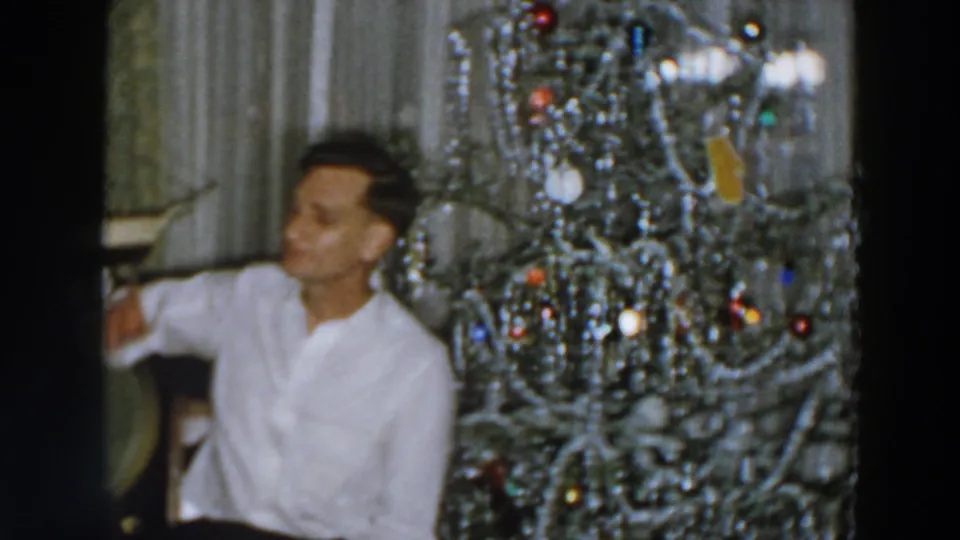 The seated man appears to speak or listen off-camera beside the tinsel Christmas tree in the 1957 Wheeling interior.