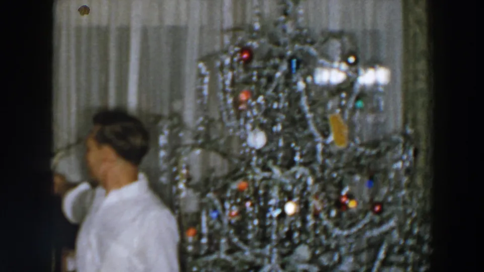 A side view shows the man leaning near the chair with the tinsel Christmas tree and ornaments filling much of the frame.