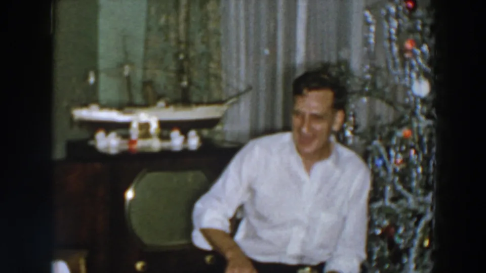 The man smiles beside the tinsel Christmas tree, with a vintage television and tabletop model ship visible behind him.