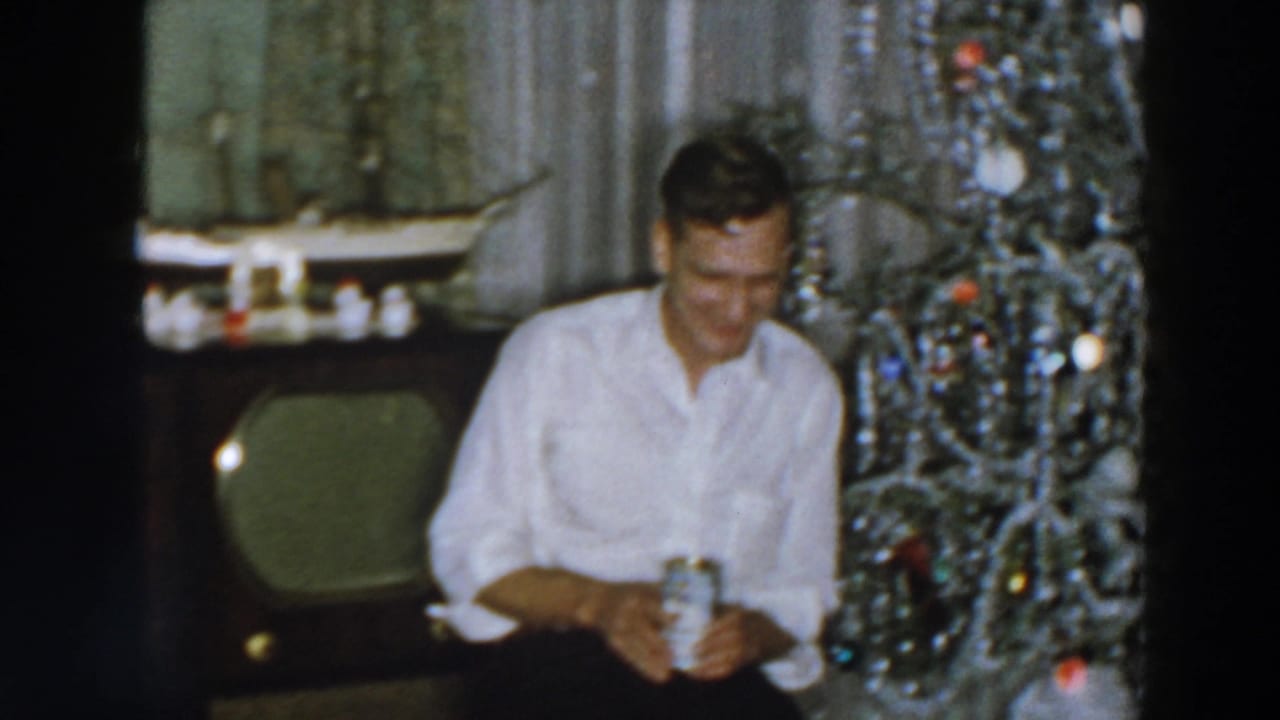 In a 1957 Wheeling, Ohio home movie frame, a man in a white shirt smiles while seated beside a tinsel-covered Christmas tree.