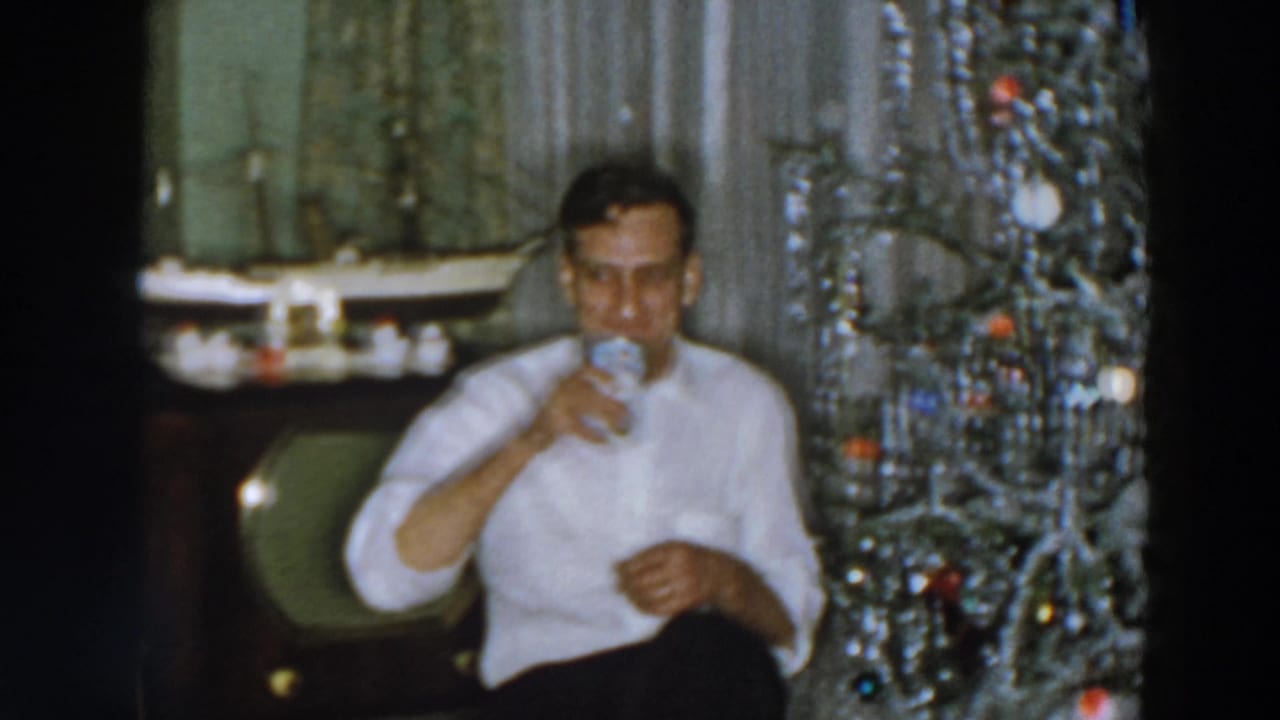 Man seated by a Christmas tree drinking from a glass.