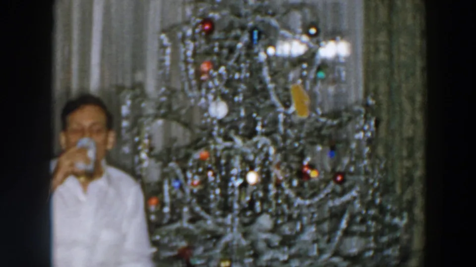 A close, off-center home-movie frame shows the man drinking beside the ornamented Christmas tree in the private holiday interior.