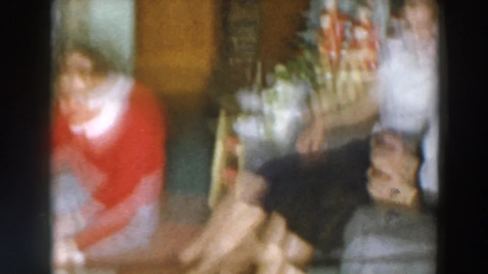 A blurred 8mm frame from the Wheeling Christmas interior shows the woman in red moving near seated adults and festive decor.