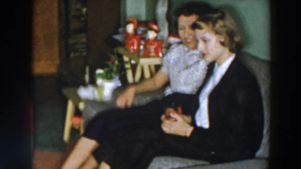 Two women sit together on a sofa in the 1957 Wheeling living room, with holiday decorations and a small reindeer figure nearby.